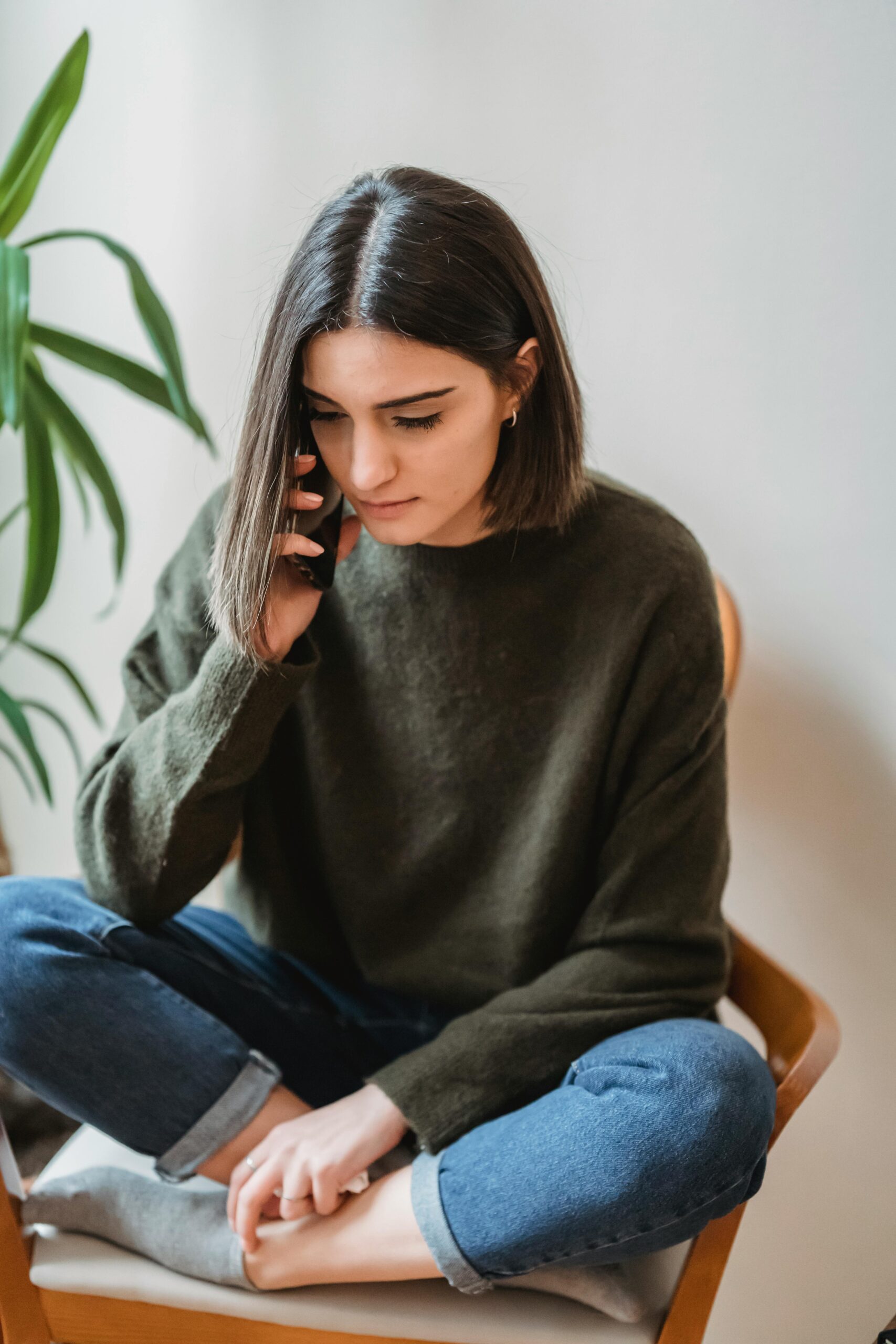 Full body of young ethnic lady with dark hair in casual outfit sitting on chair with crossed legs and talking on smartphone at home